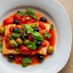 Pesce persico alla pizzaiola con pomodoro olive e capperi su piatto bianco