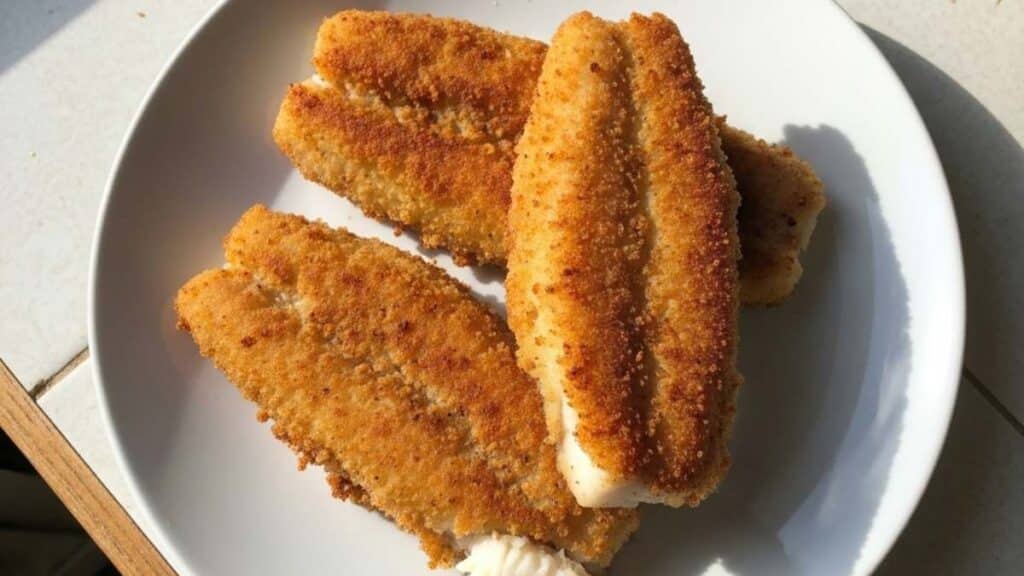 Pesce persico fritto dorato e croccante su piatto bianco con limone